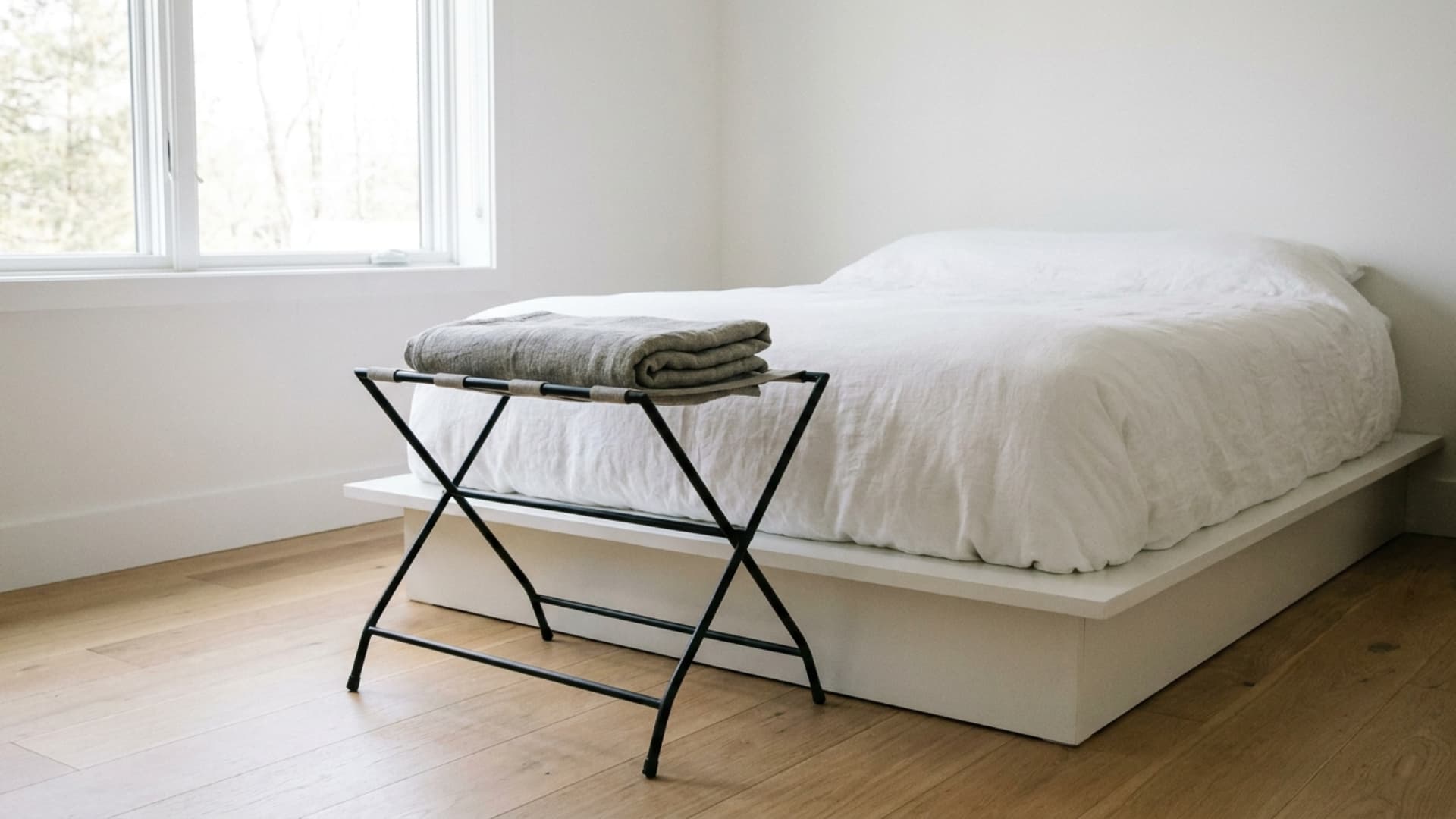 slim metal luggage rack at foot of white guest bed with folded grey linen blanket