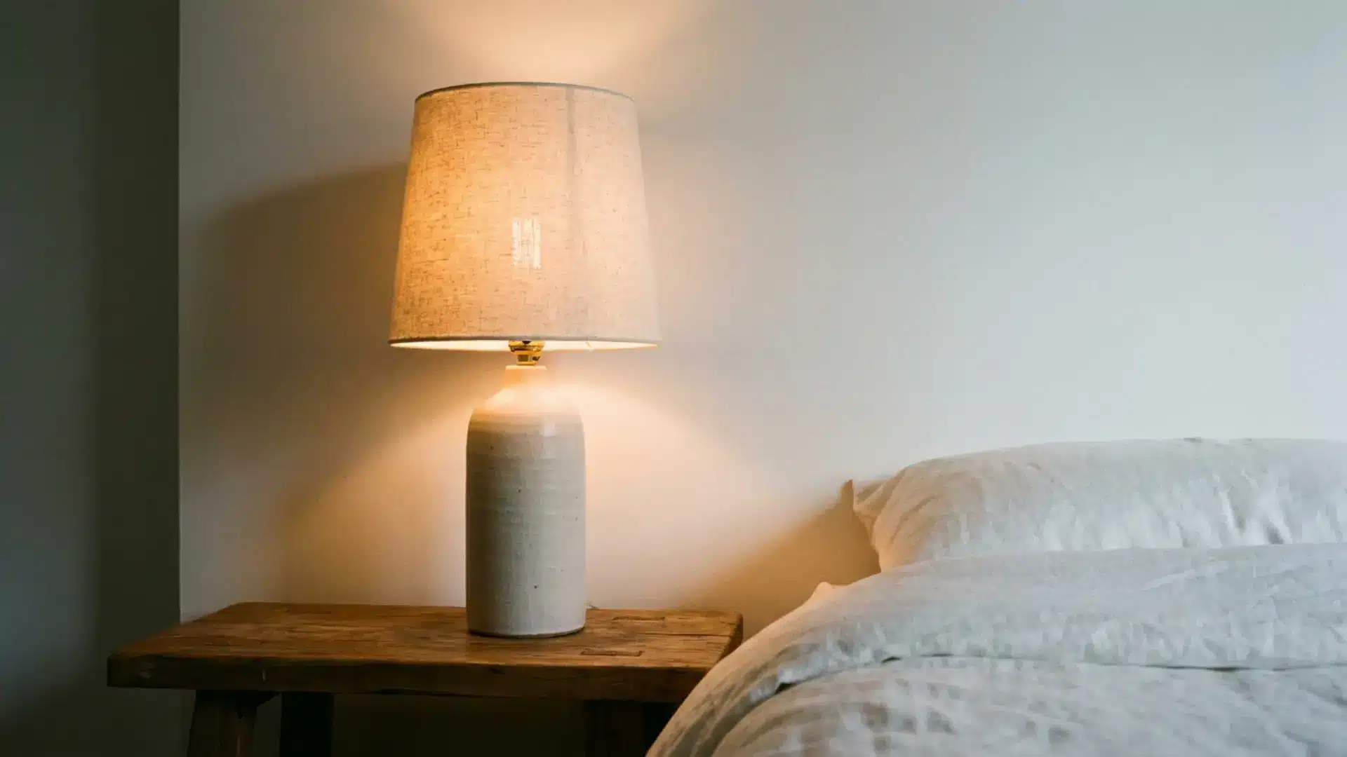 slim ceramic bedside lamp casting warm amber glow on white wall beside white guest bed