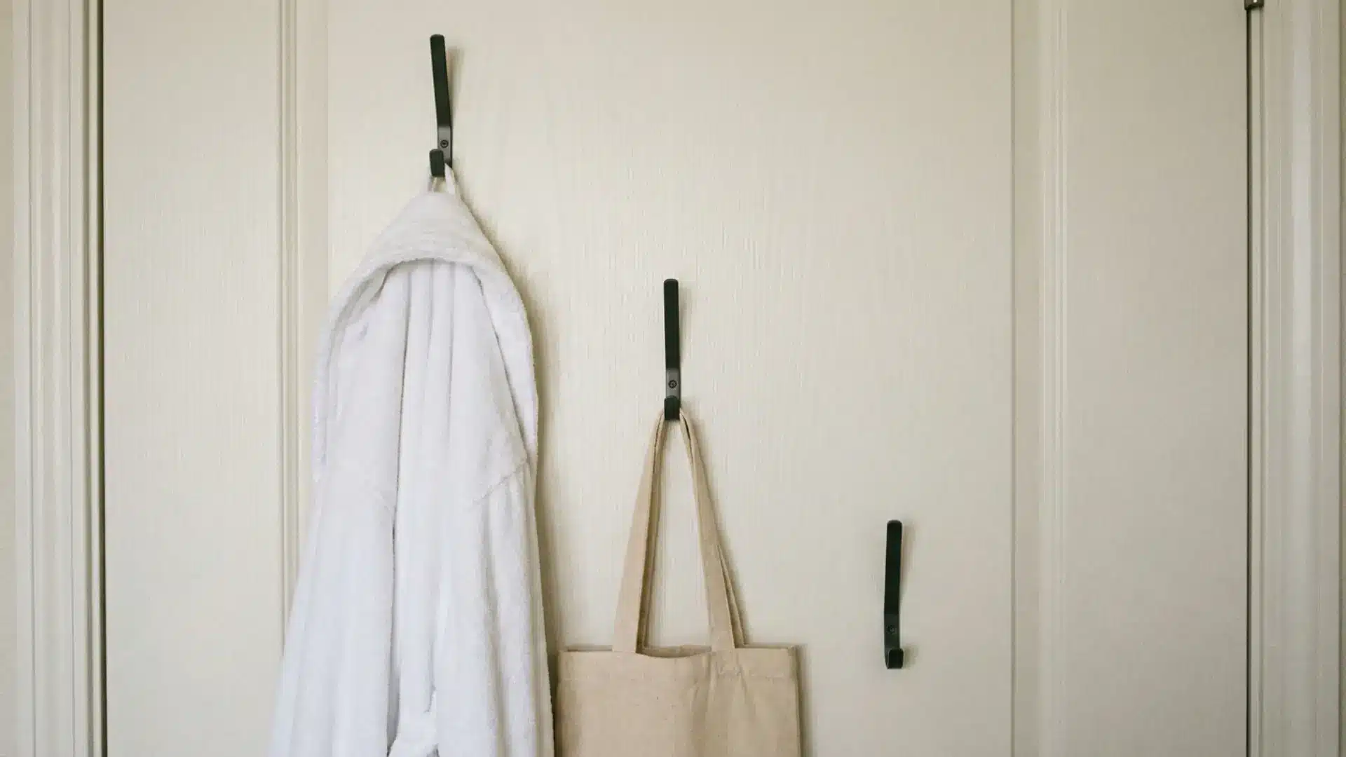 back of white guest room door with three matte black hooks holding robe and canvas tote