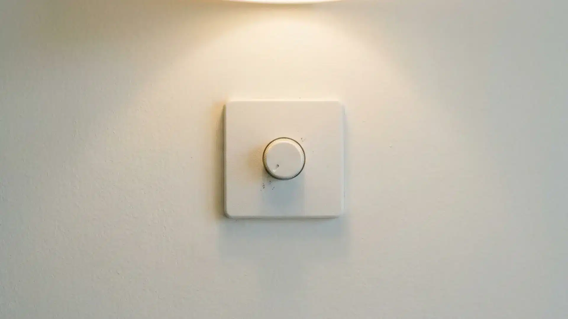 matte white dimmer switch on plain white wall with warm overhead light glow visible
