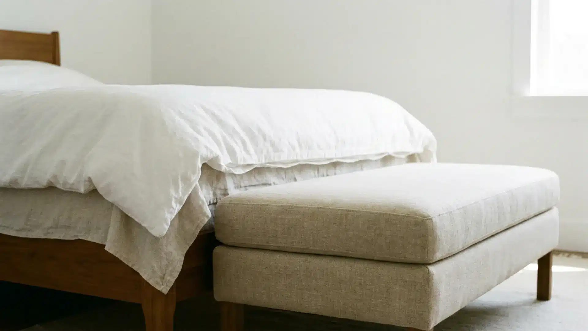 low rectangular linen ottoman at foot of white minimalist guest bed against plain wall