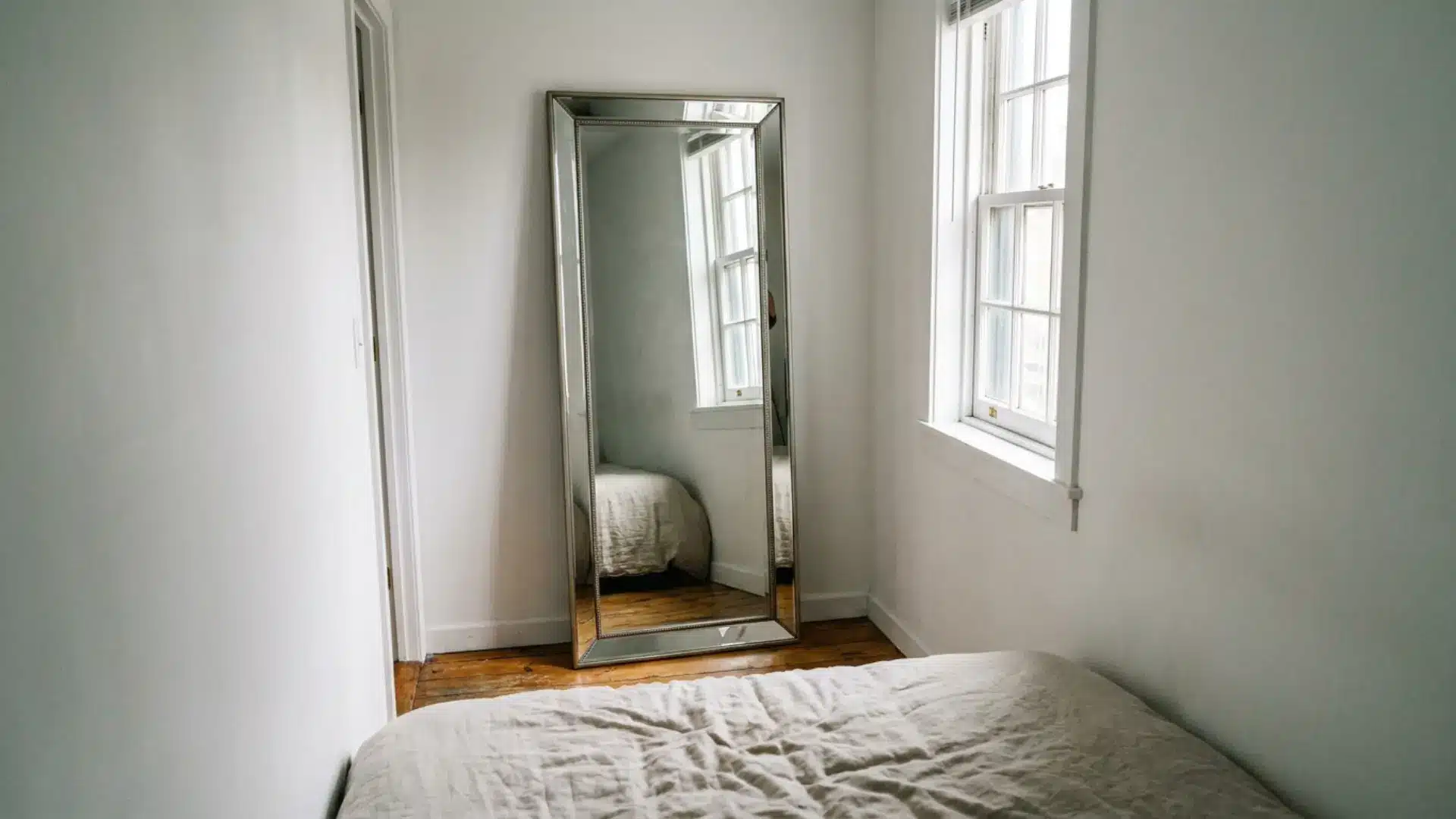 full-length mirror reflecting window light in narrow white guest room from foot of bed