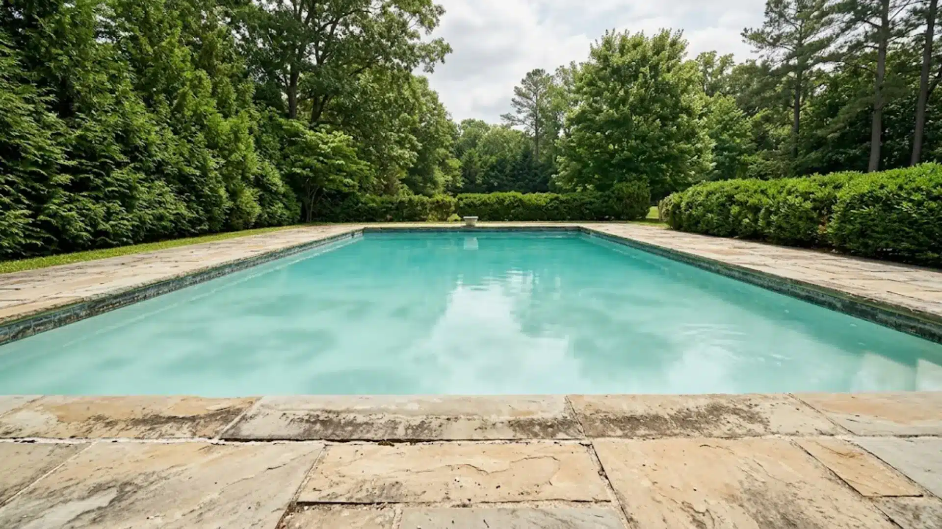 outdoor swimming pool with hazy milky turquoise water and stone deck surrounded by green hedges