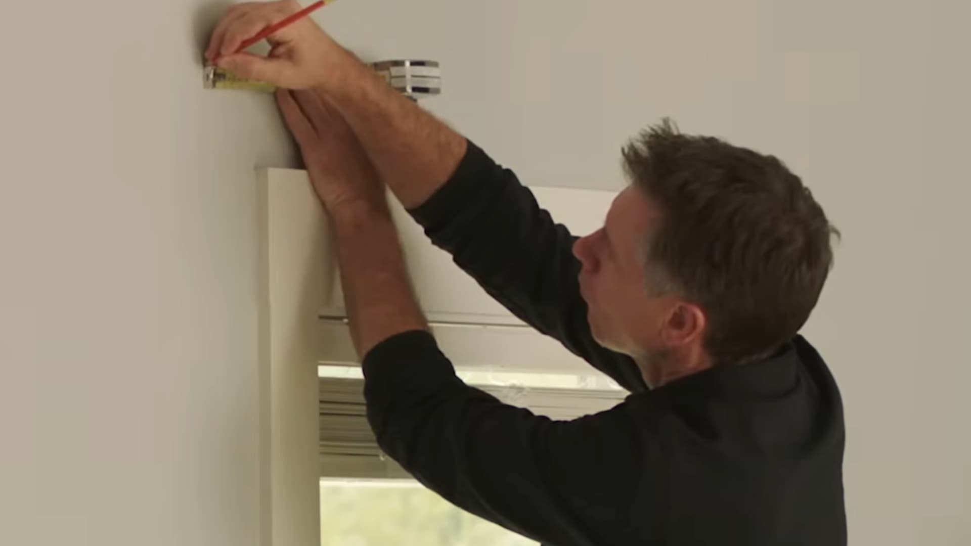 man marking bracket placement above window frame with pencil and tape measure