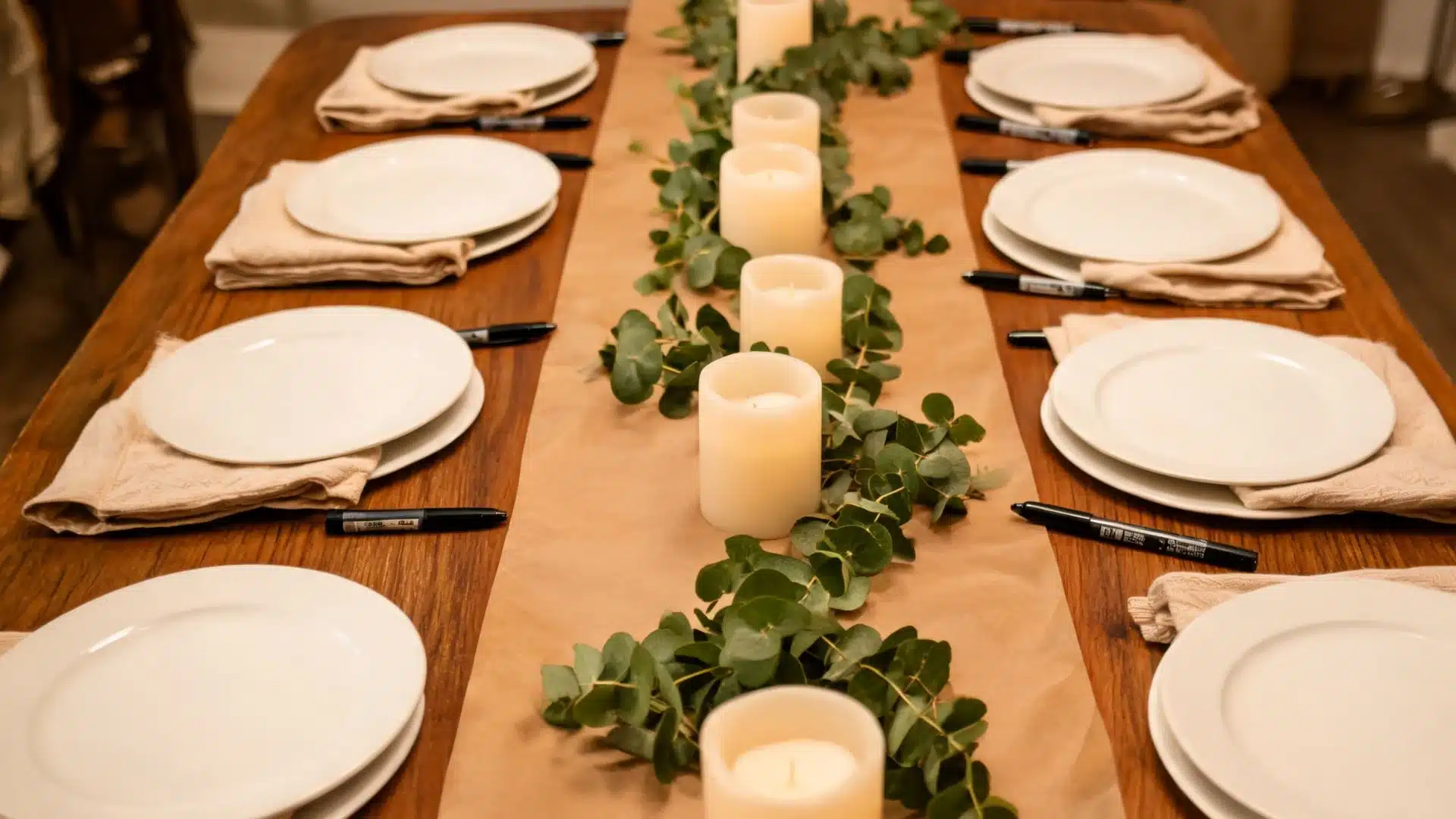 blank kraft paper runner with markers, eucalyptus garland, pillar candles, and white plates