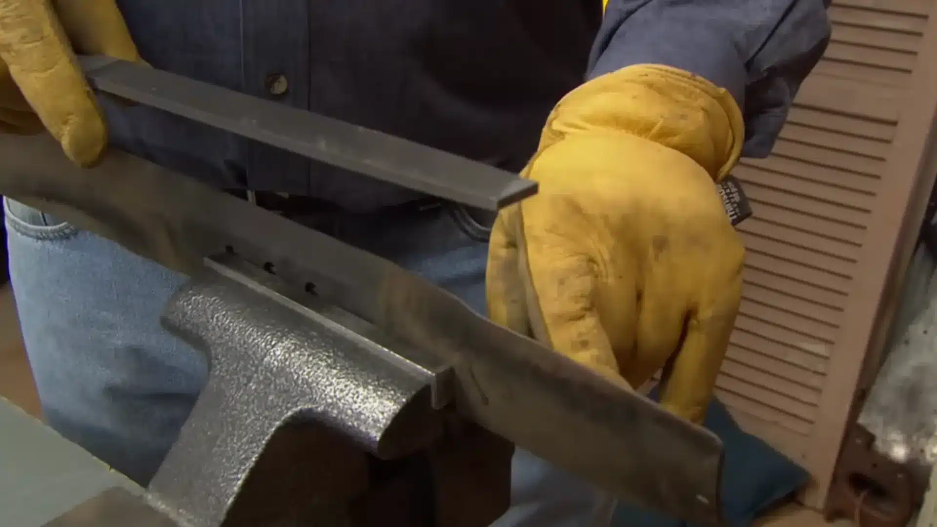 gloved hands filing a lawn mower blade's cutting edge while secured in a vise