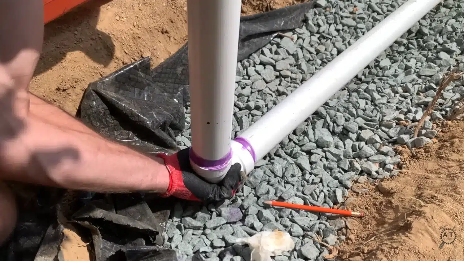 gloved hands gluing a purple primer coated pvc drain pipe fitting on a gravel bed