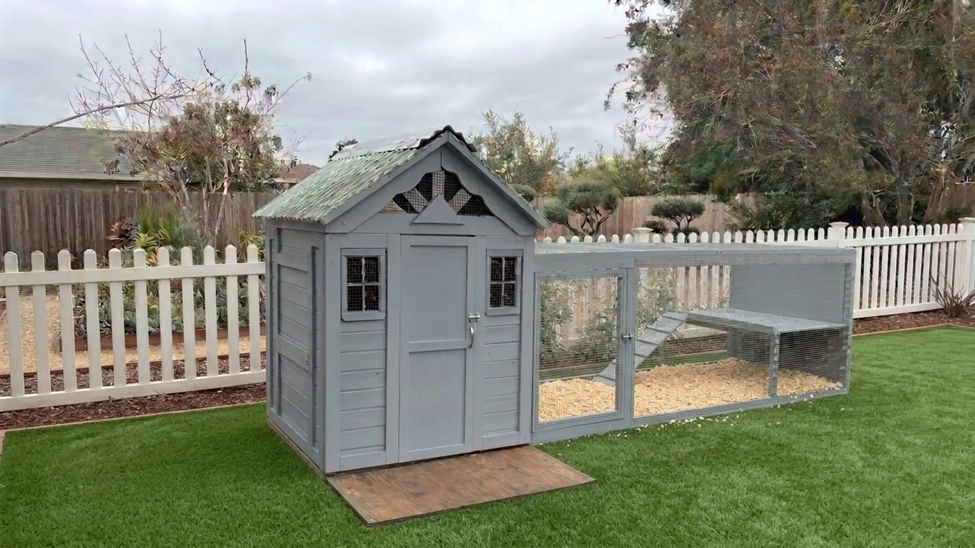 gray converted playhouse chicken coop with an attached wire run inside a white picket fenced gardens