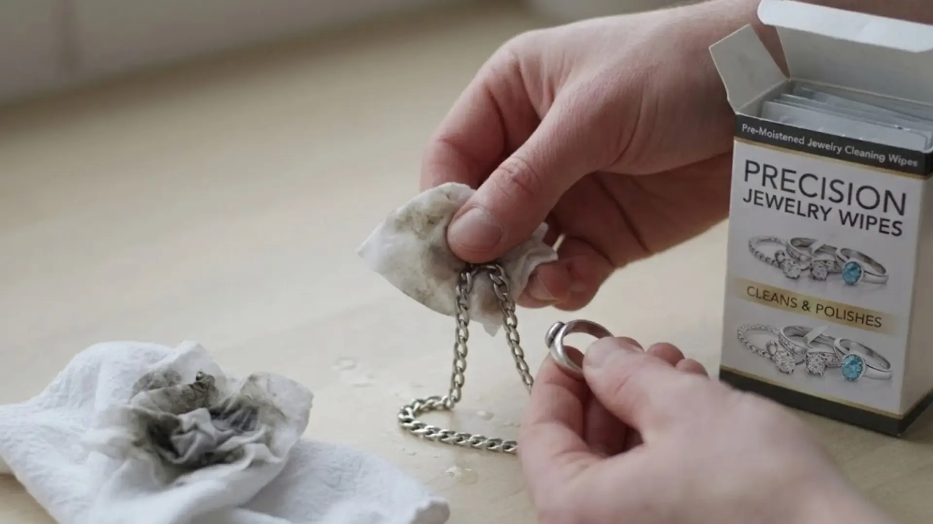 hands cleaning a silver chain and ring with jewelry wipes beside a box of precision cleaning wipes