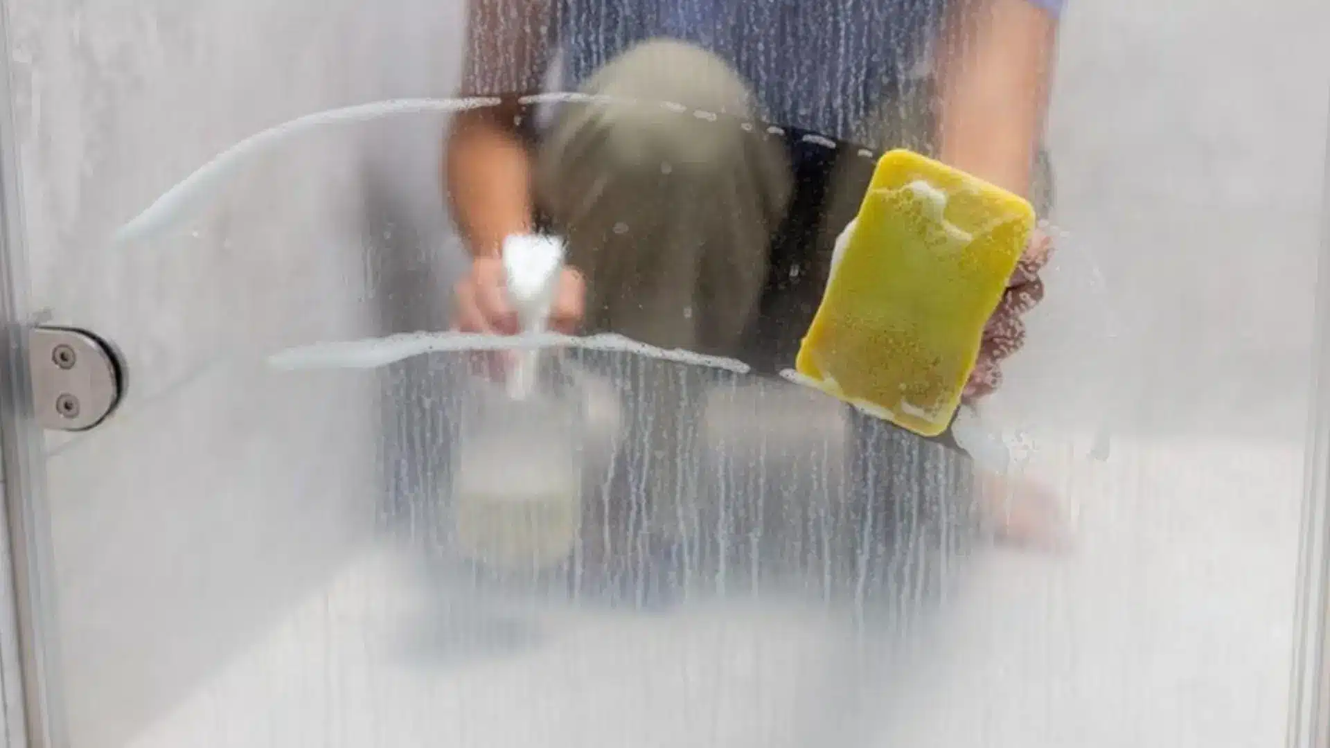 person cleaning a cloudy glass shower door with a yellow sponge and soapy water to remove mineral buildup