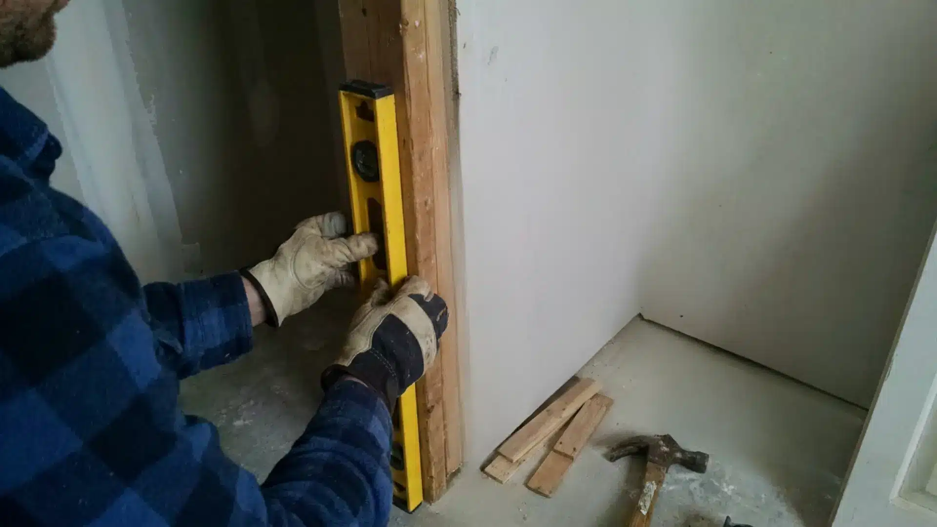 man in gloves holding a level against a door frame jamb with shims and hammer nearby