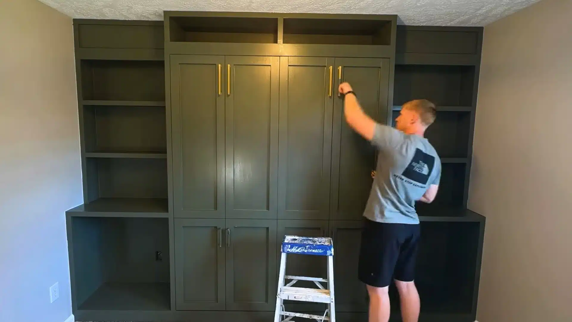 man attaching a brass drawer pull to a painted cabinet door while standing on a step ladder