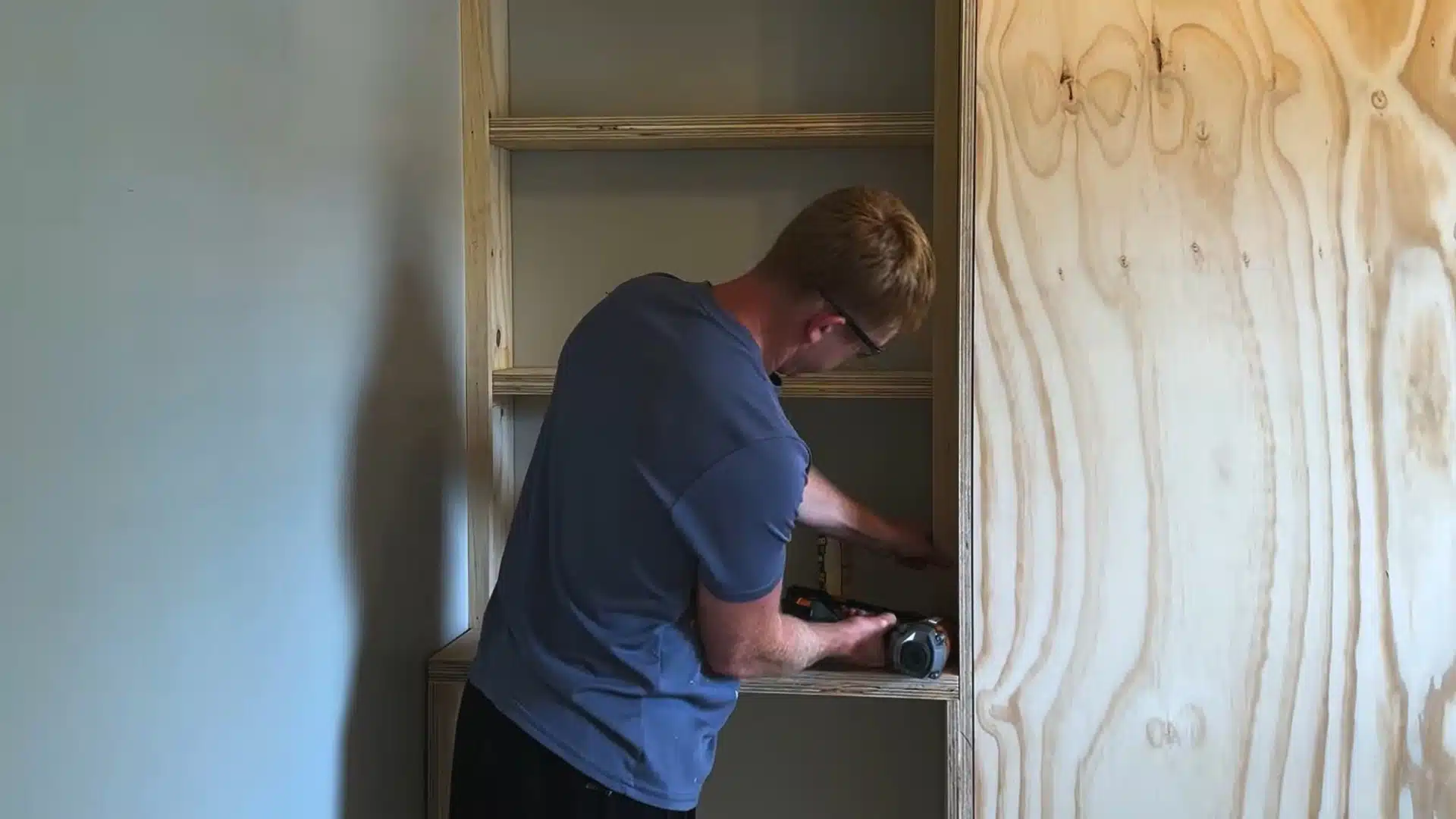 man drilling inside an open cabinet shelf frame next to a stack of plywood panels