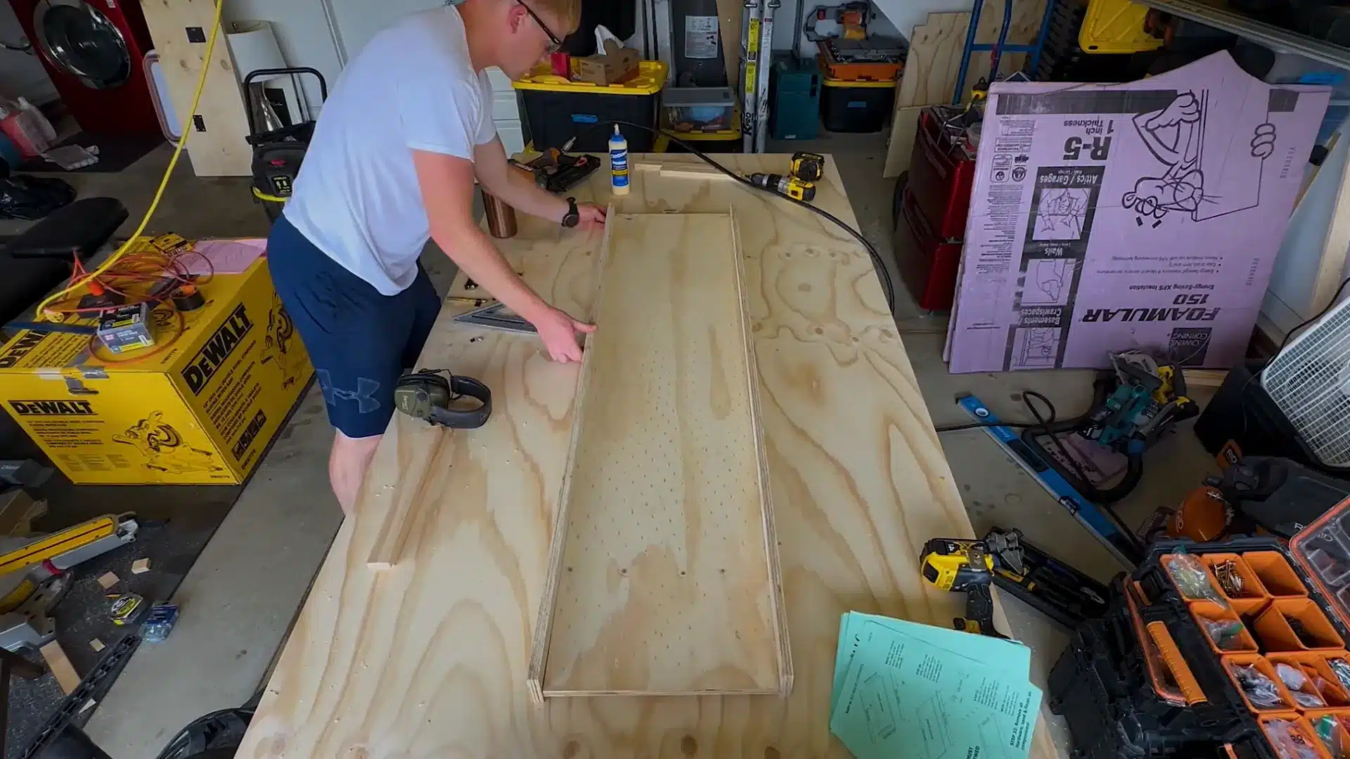 man gluing and aligning a long plywood board on a cluttered garage workbench surface