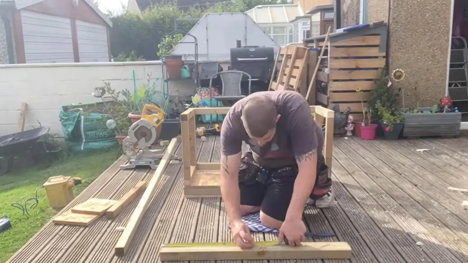 man kneeling down to measure a wooden plank near a partly built box frame on an outdoor deck