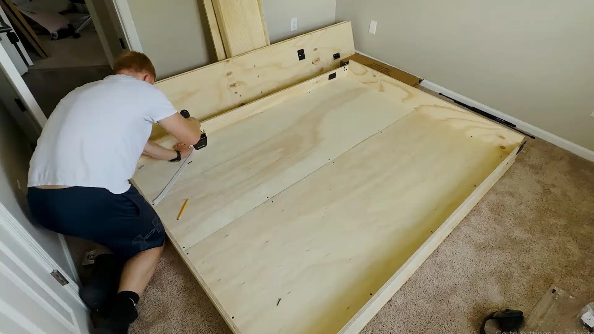 man kneeling on the carpet to drill a plywood panel onto the assembled bed frame