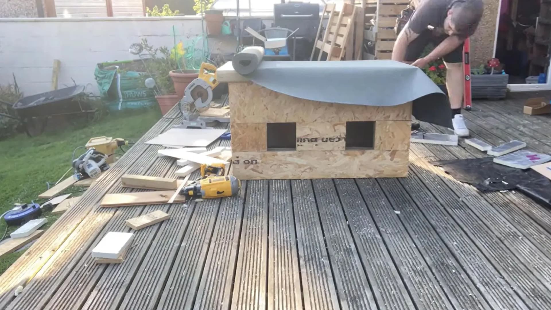 man laying grey roofing felt over the top edge of an osb cat house structure outdoors