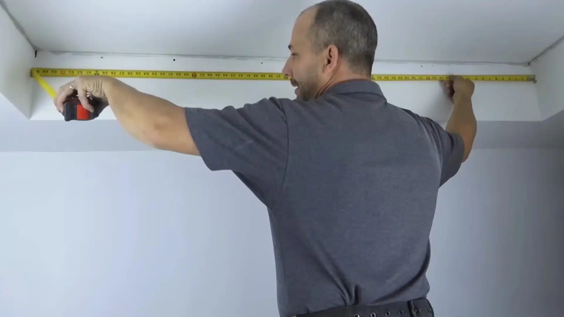 man measuring ceiling corner with tape measure before installing crown molding in a plain white room