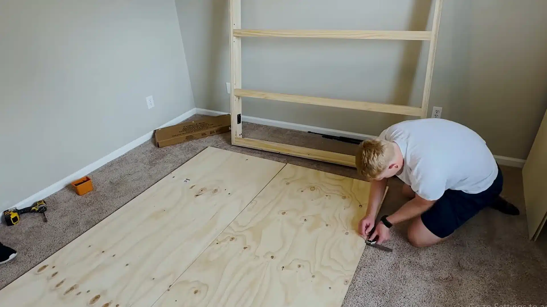 man measuring plywood panels on the carpet beside an assembled bed frame section