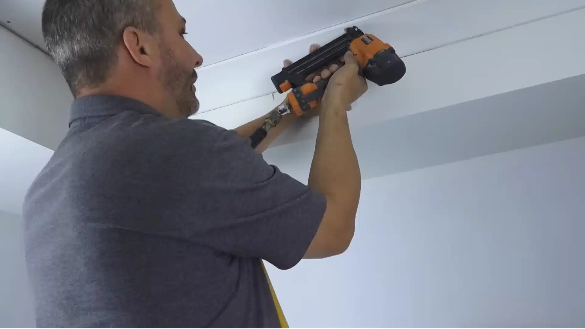 man using orange pneumatic nail gun to secure crown molding trim along ceiling and wall corner lines