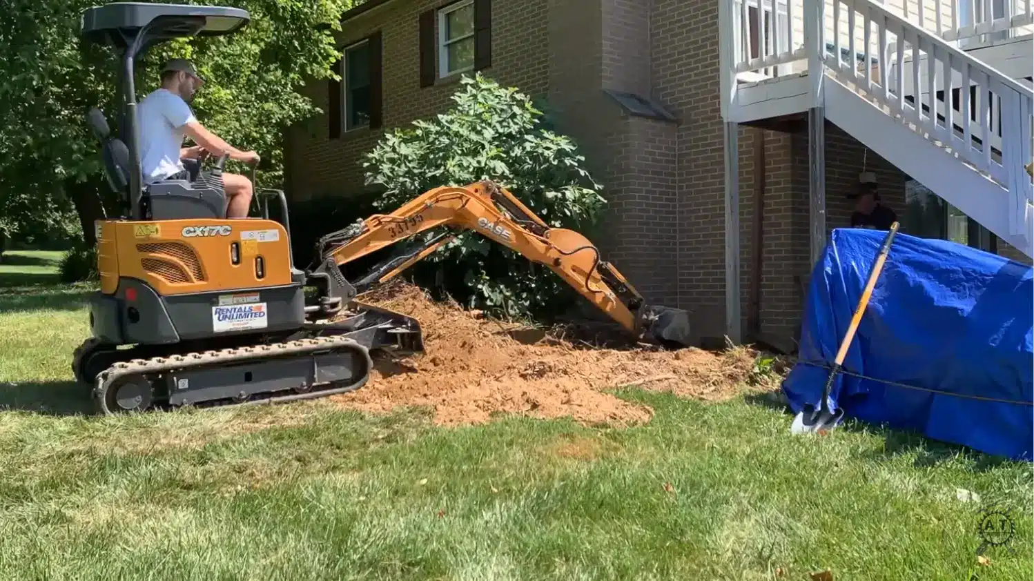 mini excavator digging out a shrub and root system near a brick house foundation