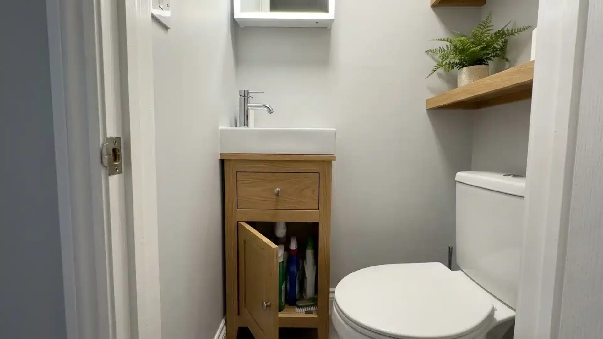 narrow wood vanity cabinet with open door showing cleaning supplies beside a toilet