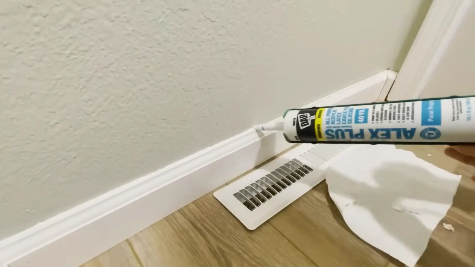 person caulking the top edge of an installed baseboard near a floor vent