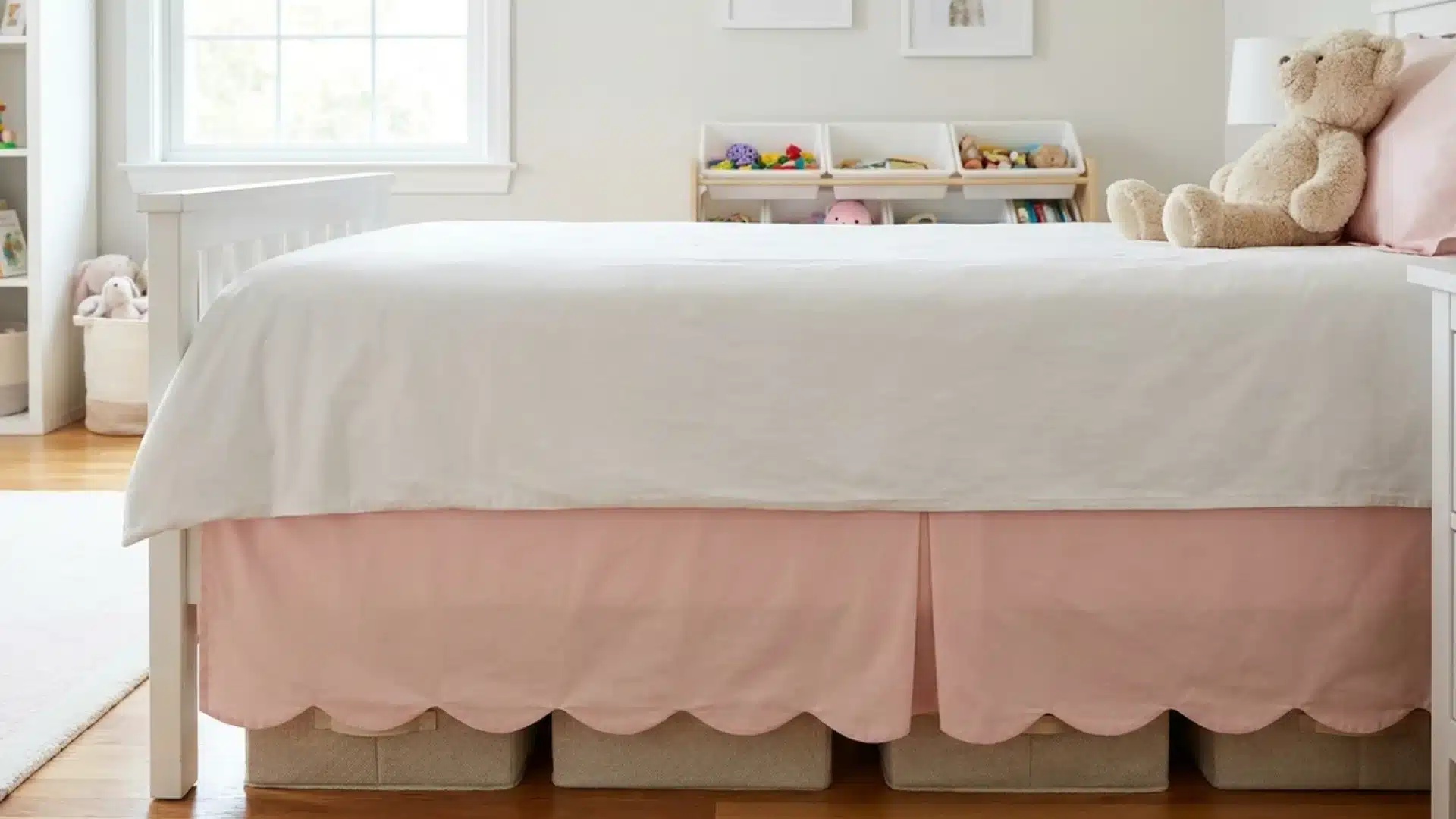pink scalloped bed skirt covering storage bins beneath a white bed with a teddy bear