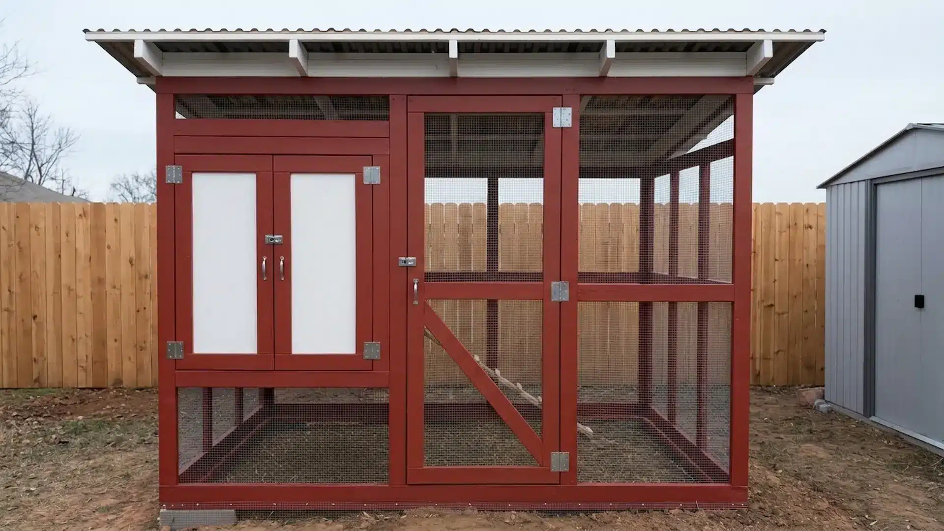 red compact chicken coop with a wire mesh run set against a tall wooden fence near an urban backyard