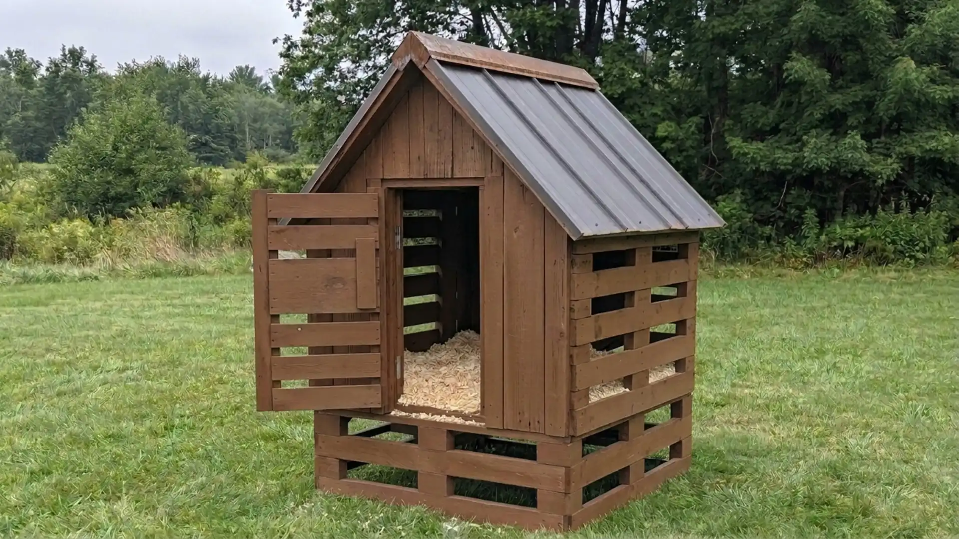 rustic chicken coop built from wooden pallets with a metal roof standing in an open grassy field now