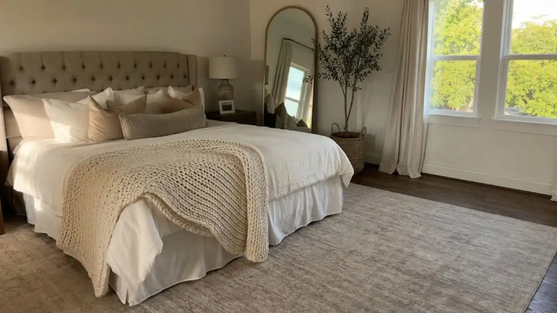tufted taupe headboard with cream bedding, chunky knit throw, and arched floor mirror