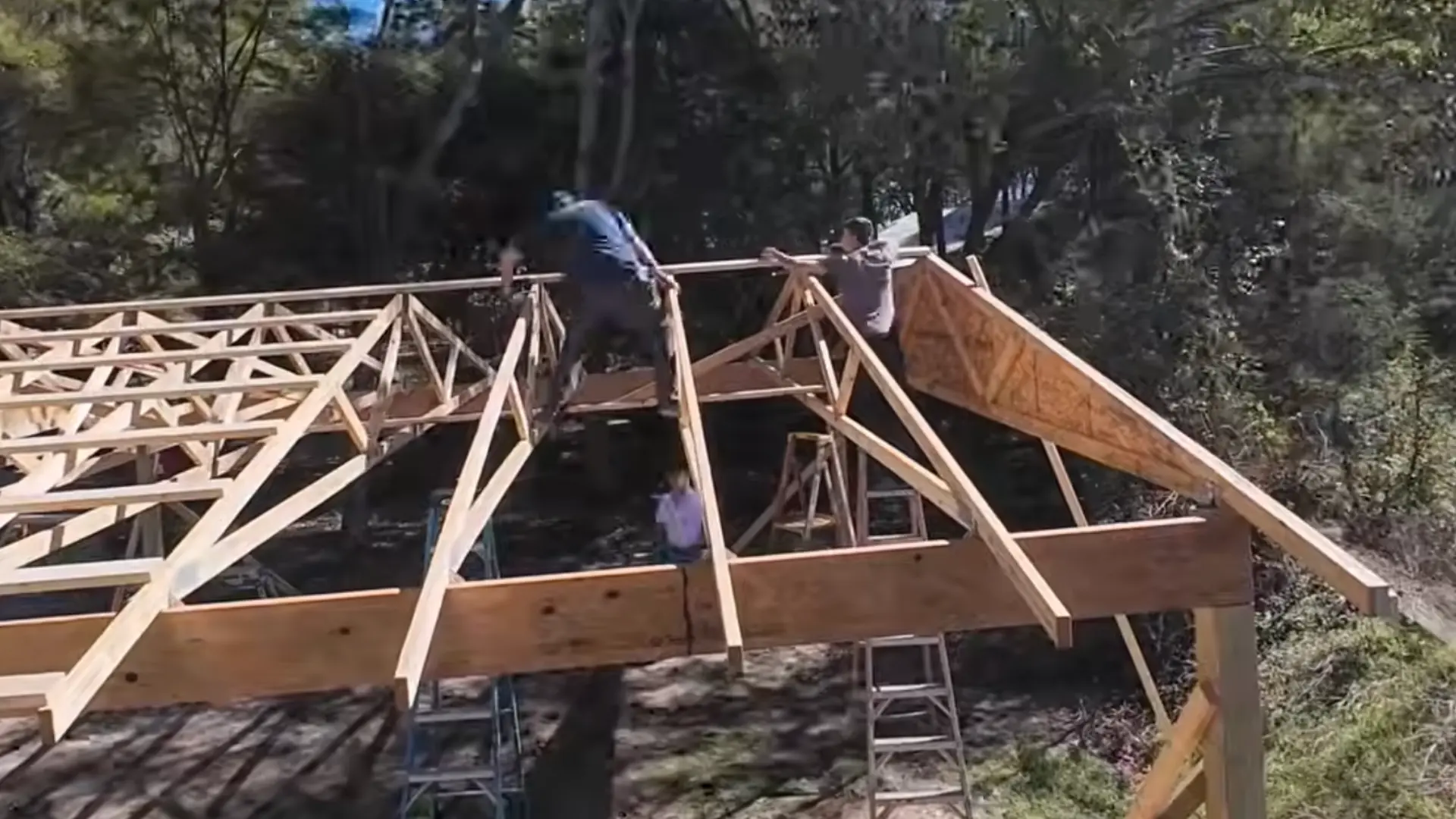 two workers carrying a long wooden board across sawhorses at an outdoor construction site