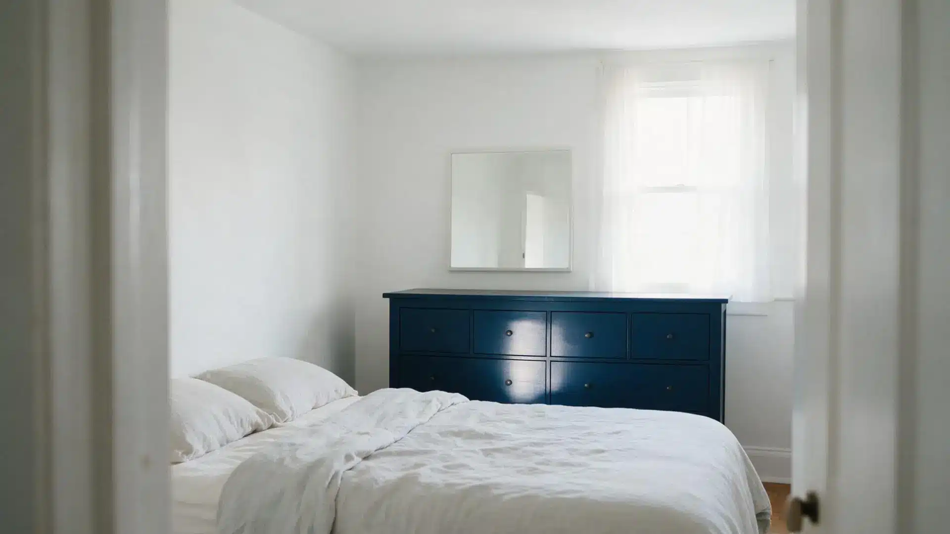 navy blue dresser with square mirror in a white bedroom seen through an open doorway and sheer curtains at the window