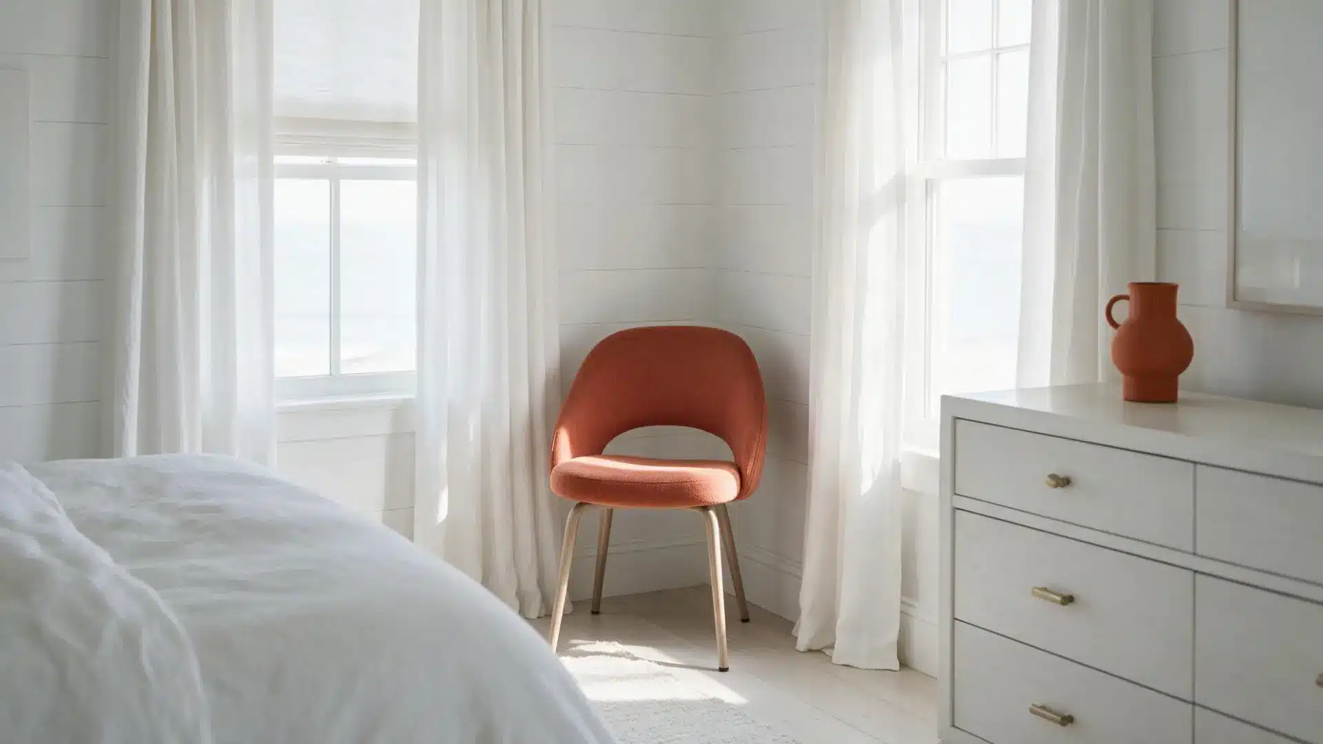 terracotta accent chair in the corner of a white coastal bedroom near sheer curtains and a terracotta pitcher nearby