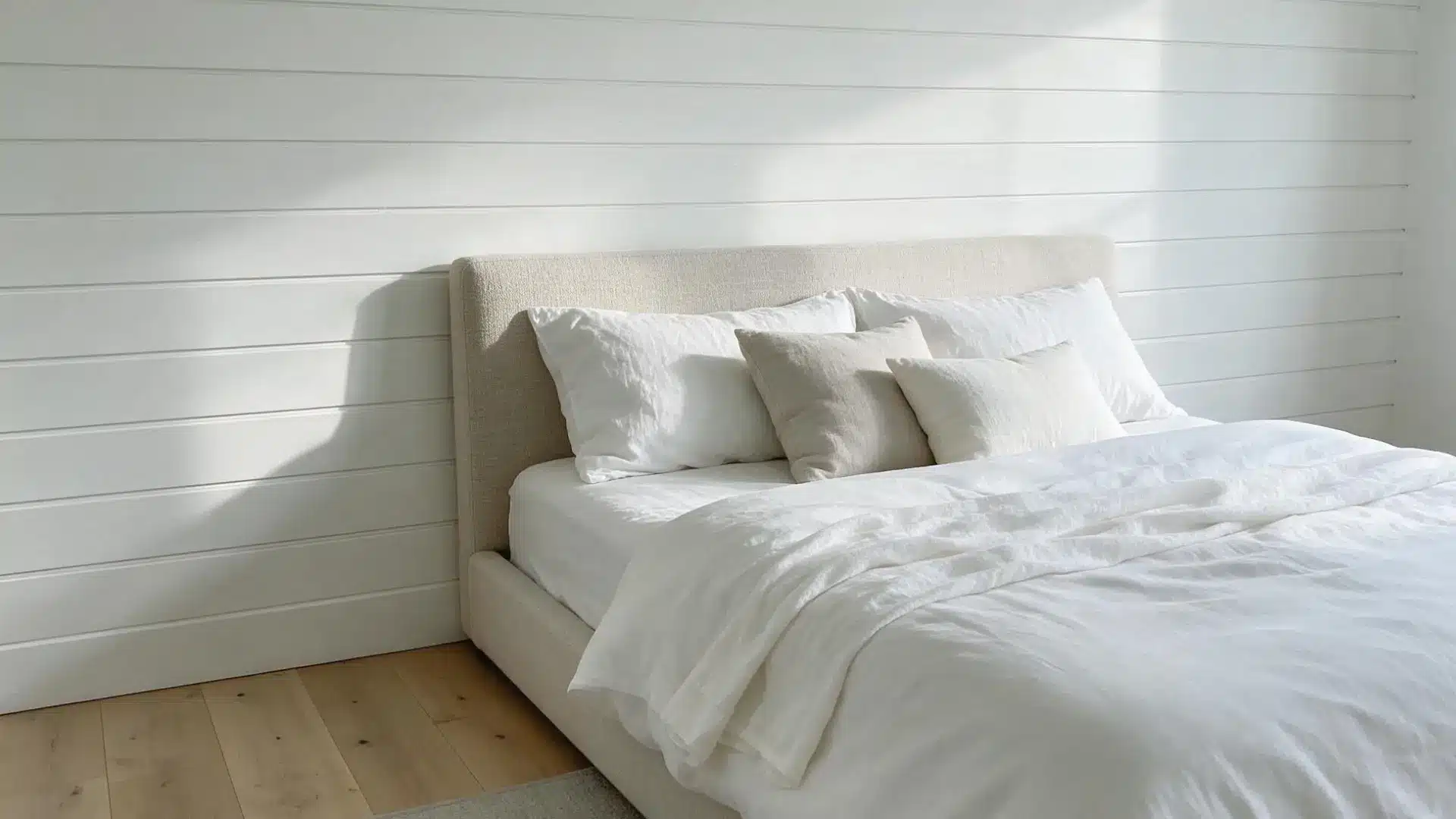 white shiplap accent wall behind a beige upholstered headboard with soft white linen bedding and light wood flooring